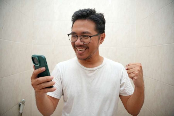 Homme souriant en regardant son téléphone, croyant avoir trouvé une bonne affaire malgré les fausses promotions lors des soldes.