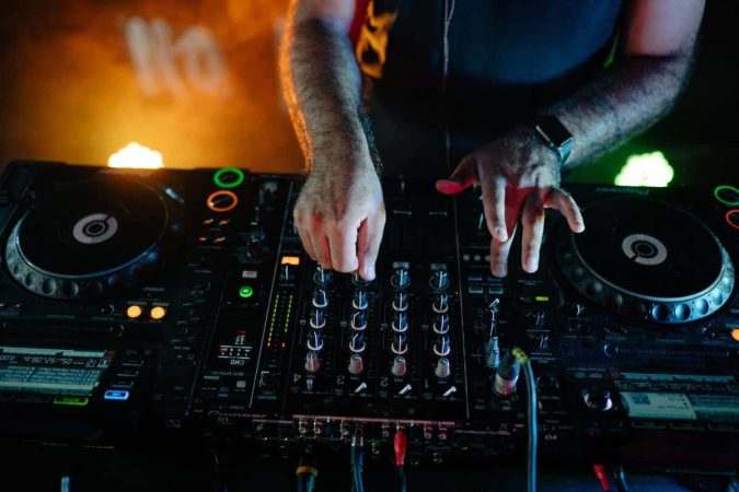 Gros plan sur les mains d'un dj mixant de la musique sur une platine Pioneer dans un club sombre avec des lumières colorées.