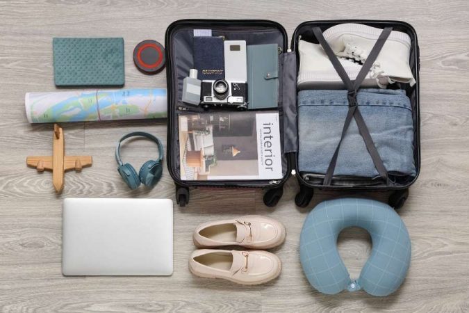 Une valise ouverte contenant des objets de voyage essentiels comme un appareil photo, un casque audio, des chaussures et un coussin de cou.