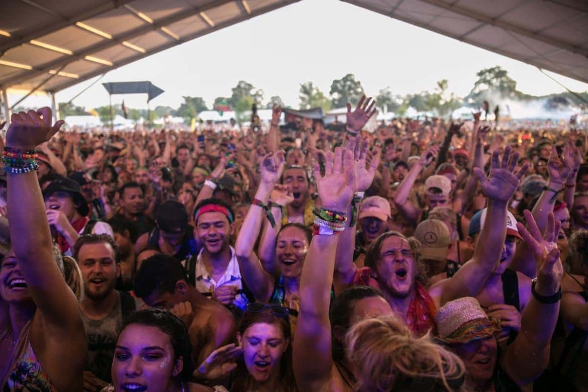 Une foule immense et enthousiaste lève les mains en l'air lors d'un festival de musique électro sous un grand chapiteau en plein air.