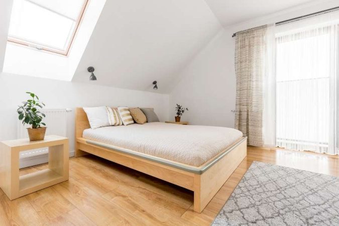 Un lit au sol 2 places en bois clair, équipé de coussins décoratifs, dans une chambre mansardée lumineuse avec des plantes et un velux.