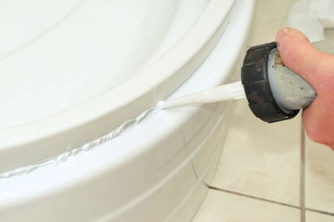 Une main applique des joints en silicone le long d'une baignoire blanche pour assurer l'étanchéité.
